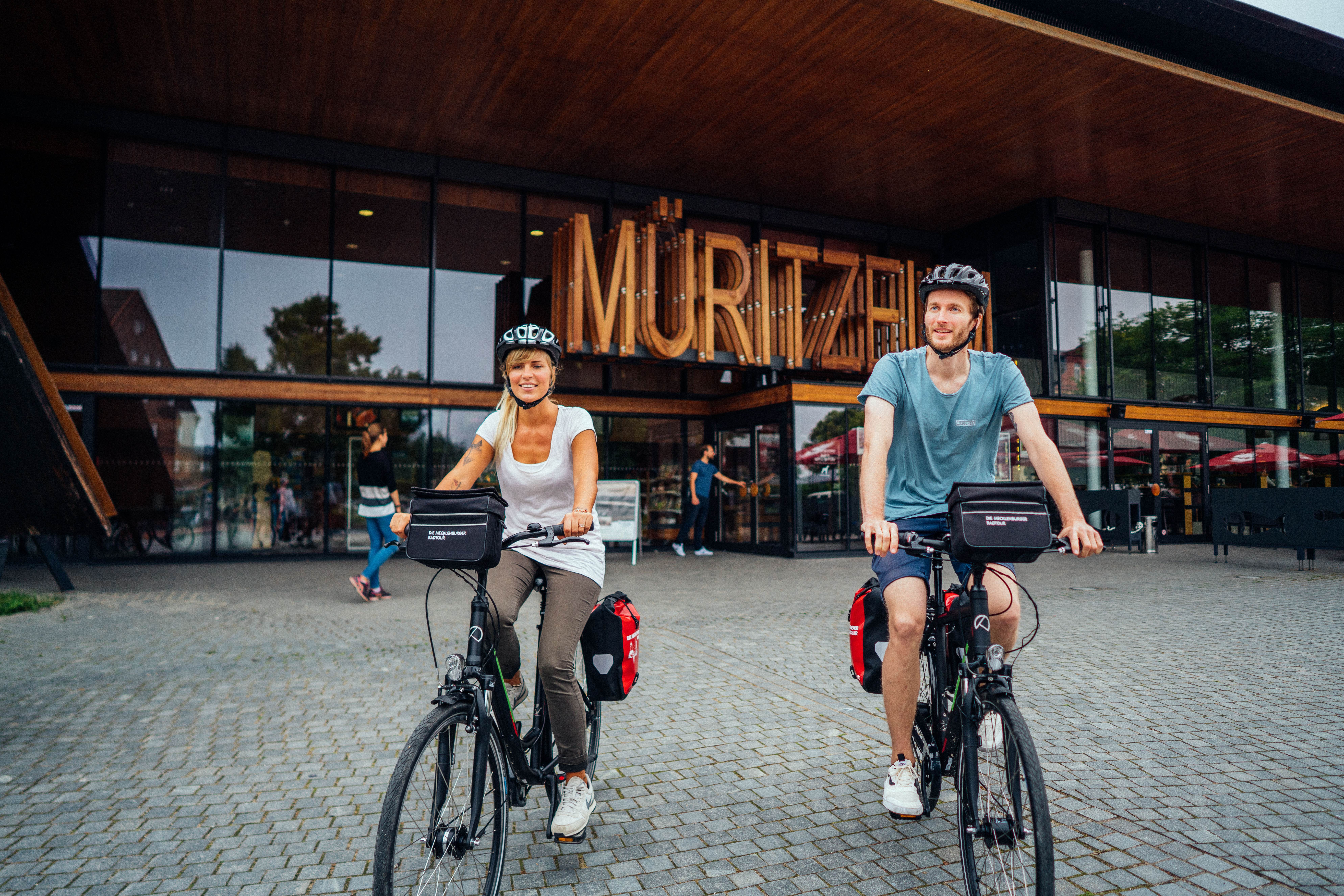Ein paar startet seine Fahrrdtour vom Müritzeum aus. Beide schauen lächelnd in die Kamera, im Hintergrund sieht man den Eingang vom Müritzeum